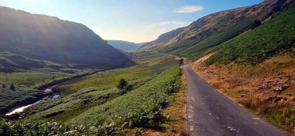 Single-track lane winding through wide green valley between steep hillsides with stream running along left edge under bright late afternoon sun.