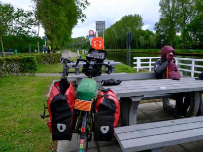 A stop alongside a canal in Belgium. We had a coffee here. Touring bike with panniers parked by a canal‑side picnic table where a person sits.