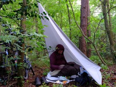 Dave in his bivy at the pitch we made on day one Person in bivy bag under a white tarp shelter with bikes in dense woodland.