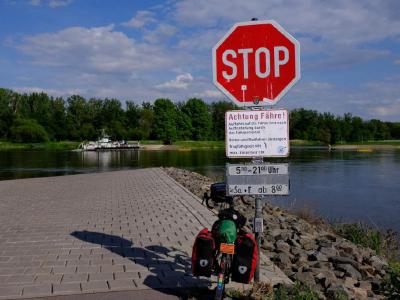 Waiting for a ferry in the Netherlands. The operator was full of chat and cheer. Stop sign and ferry warning above a loaded touring bike at river slipway.