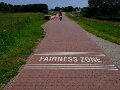The start/end of a 'Fairness Zone' near Kempen in Germany. A section of a shared path where walkers and cyclists are reminded to behave considerately, slow down, and share space safely. Cyclist on red‑paved path marked “FAIRNESS ZONE” between grassy fields.