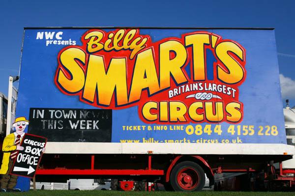 A large circus advertisement billboard on the side of a truck or trailer. The colorful sign reads &ldquo;Billy Smart&rsquo;s Circus&rdquo; with &ldquo;Britain&rsquo;s Largest Circus&rdquo; underneath, advertising that it&rsquo;s &ldquo;In Town This Week&rdquo; with ticket and contact information displayed.