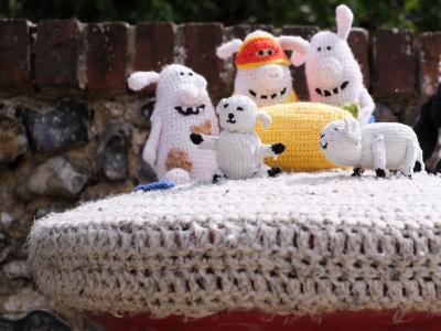 A crocheted postbox topper featuring a group of knitted characters from Shaun the Sheep, including Shaun, Bitzer the dog, and the flock, standing on a light-coloured base atop a red Royal Mail postbox with a brick wall in the background.