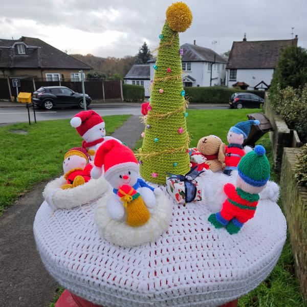 A festive knitted Christmas scene displayed on top of a red Royal Mail post box in a residential street. The handcrafted topper features a winter wonderland tableau on a circular white crocheted base with red detail stitching. At the center stands a tall green spiral Christmas tree decorated with colorful baubles in pink, blue, and other bright colors, topped with a golden yellow star. Surrounding the tree are multiple knitted figures: two white snowmen wearing red Santa hats with white pompoms, one holding a yellow gift, and several small dolls dressed in colorful winter clothing - including figures in red, blue, and green striped outfits with knitted hats. There&rsquo;s also a brown teddy bear character among the group. The scene is photographed outdoors with suburban houses, parked cars, hedges, and a cloudy sky visible in the background, creating a charming community Christmas display.