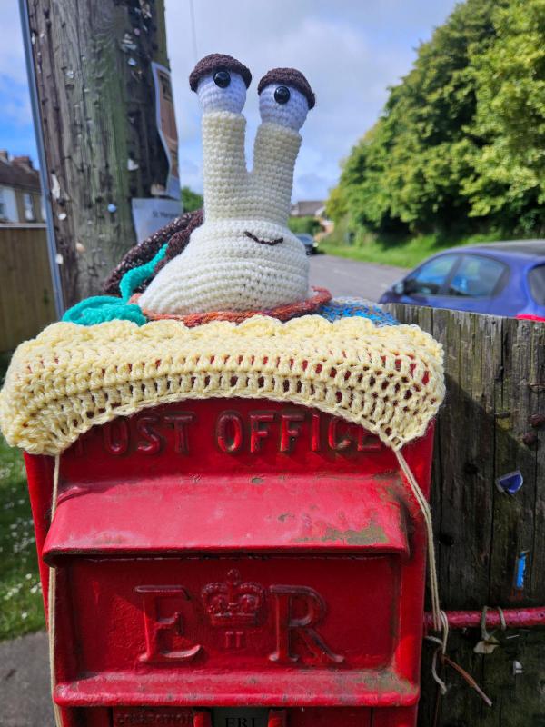 Red Royal Mail post box decorated with yarn bombing. A pale yellow crochet trim covers the ‘Post Office’ lettering on the box door. A large white crochet snail with tall stalk eyes and brown eyebrows perches on top. Residential street visible in background.