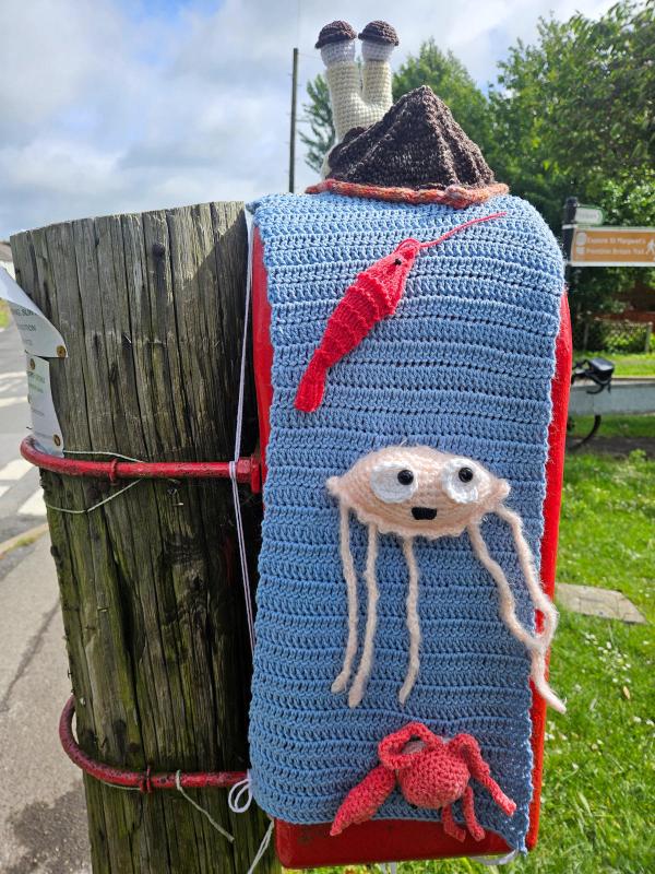 Red Royal Mail post box decorated with yarn bombing. A pale yellow crochet trim covers the ‘Post Office’ lettering on the box door. A large white crochet snail with tall stalk eyes and brown eyebrows perches on top. Residential street visible in background.
