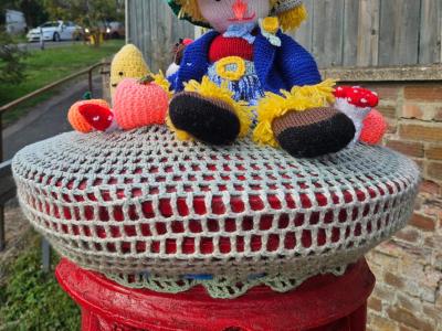 Knitted topper scarecrow sits in his green hat and blue jacket, with toy pumpkins and a mushroom beside him, edged with scalloped lace trim.