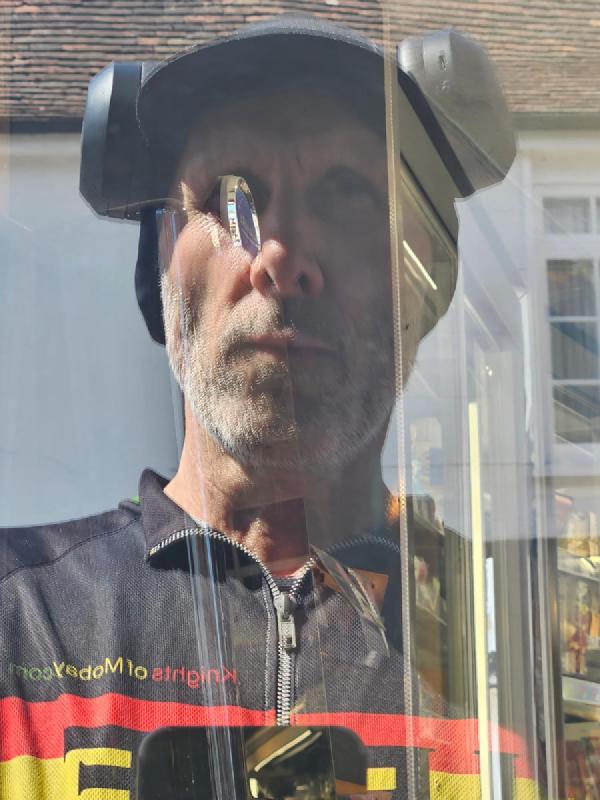 Reflection in shop window shows a cyclist wearing helmet and cap with beard, dressed in black jersey with red-yellow-white stripes, photographed through glass with strong sunlight glare. Me looking tired.