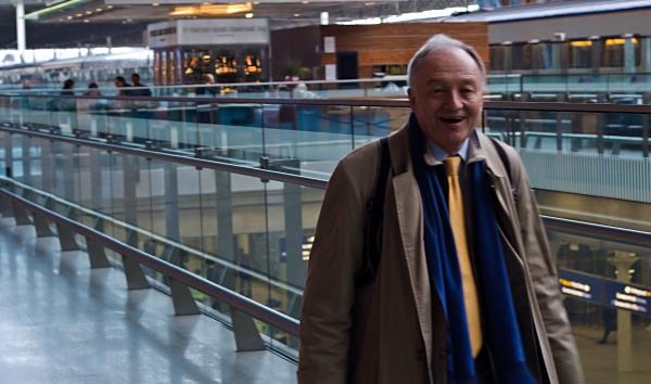 Ken Livingstone in business attire (suit jacket and tie) walking through St Pancras Station concourse, with glass railings, and contemporary architecture. He&rsquo;s carrying an umbrella.