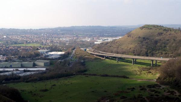 A panoramic photograph taken from high on the chalk downs above Folkestone, Kent, looking north-west across the town. In the foreground, a wide green valley floor is flanked by steep chalk and scrub hillsides. A multi-span concrete road viaduct carrying the A20/M20 approach road cuts diagonally across the valley, supported on tall concrete pillars. To the left, light industrial and warehouse buildings sit alongside a straight road leading into the town center. Beyond, rows of red-brick suburban housing spread across the middle distance. In the background, further chalk hills and the wider Kent landscape fade into a pale winter haze. There are bare deciduous trees and dormant scrub vegetation typical in early spring.