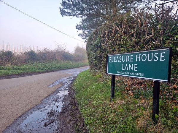A green street sign reading &ldquo;Pleasure House Lane, Borough of Maidstone&rdquo; mounted on black posts beside a narrow, wet rural lane disappearing into misty farmland, with muddy verges and hedgerows on a damp winter morning