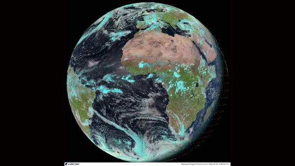 Natural-colour Meteosat satellite image of Earth centred on Africa and western Europe, with tan Sahara desert, green equatorial regions, turquoise cloud fields, and swirling weather systems over the Atlantic against a black background.